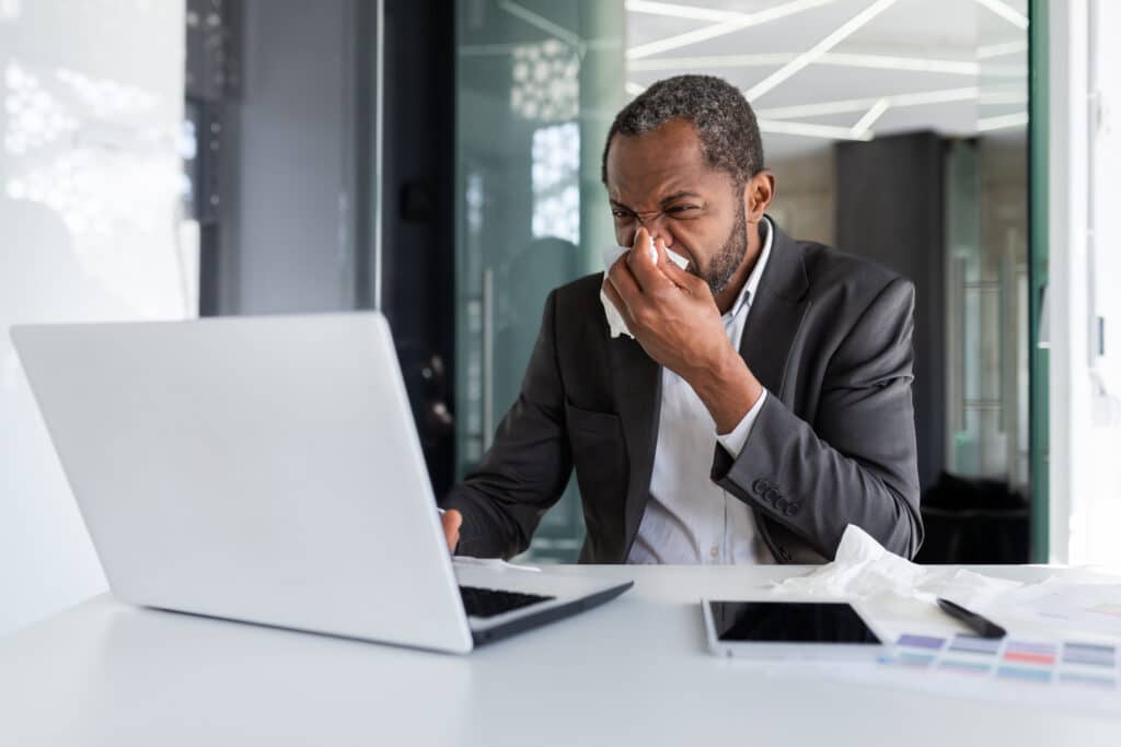 Sick man at workplace inside office with joy and cold, businessman sneezes sitting at desk, african american man with cold uses laptop at work
