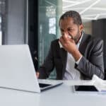 Sick man at workplace inside office with joy and cold, businessman sneezes sitting at desk, african american man with cold uses laptop at work